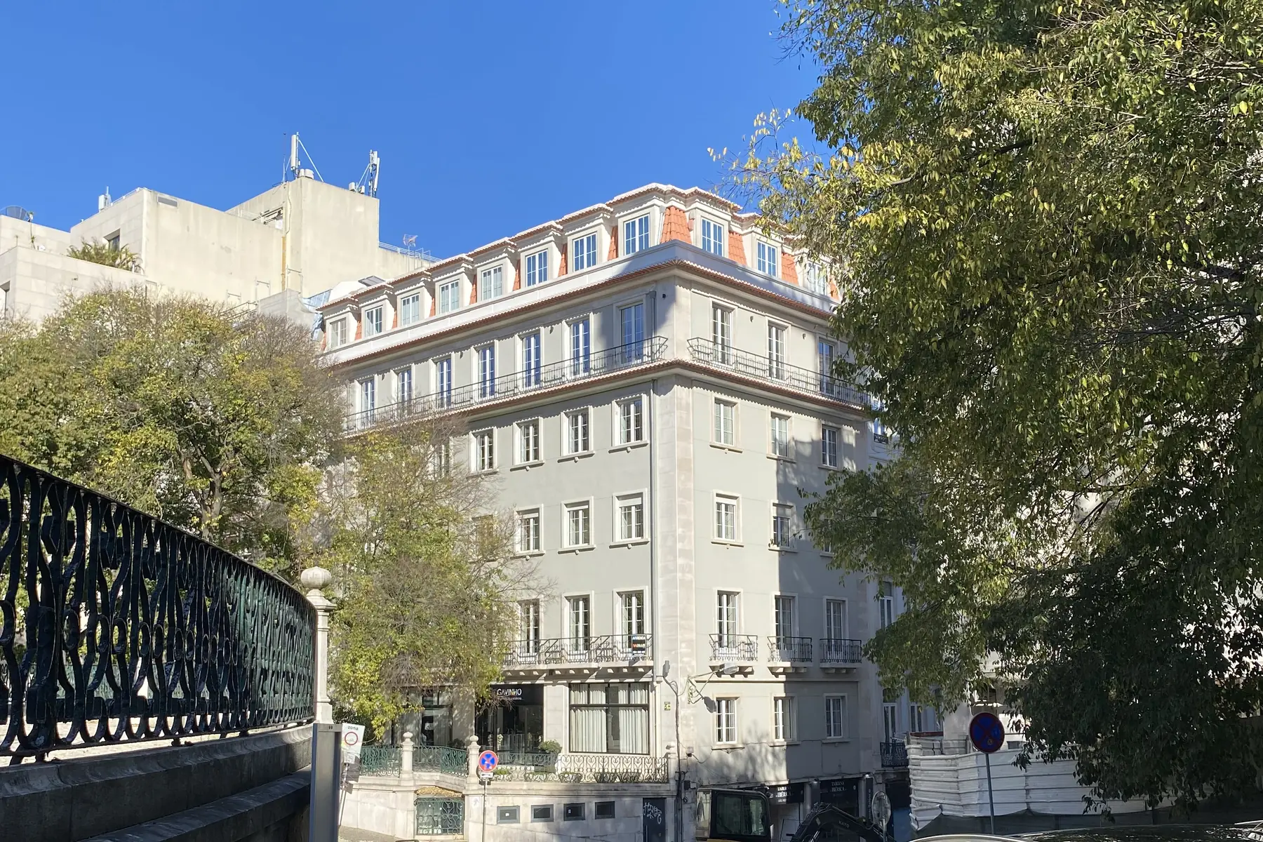 Edifício de habitação de cinco pisos, fachada clara com varandas em ferro, entre as árvores de uma rua